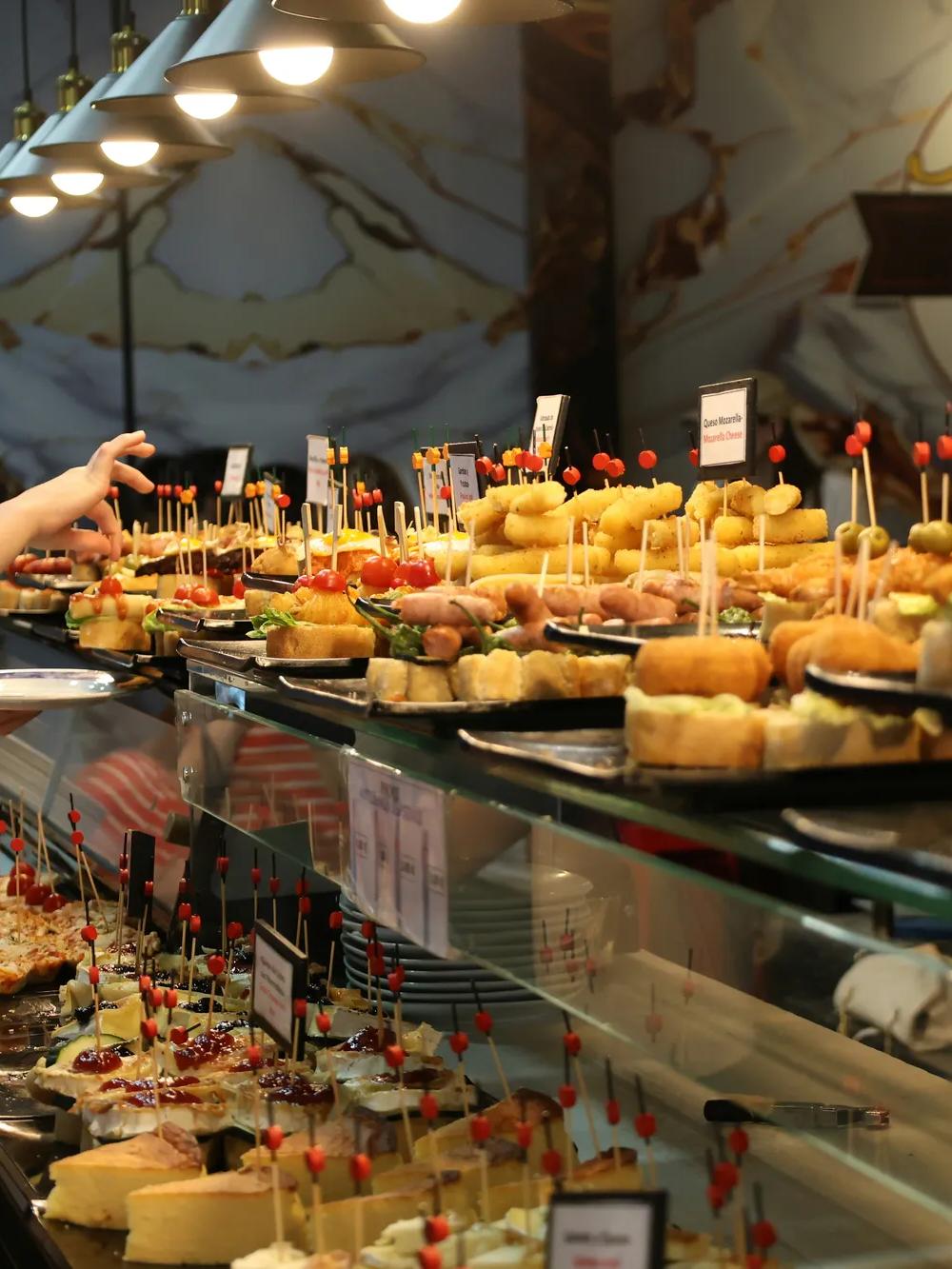 A variety of tapas and pintxos served on plates with toothpicks are displayed on a glass counter at a busy restaurant or bar, with people reaching for food.