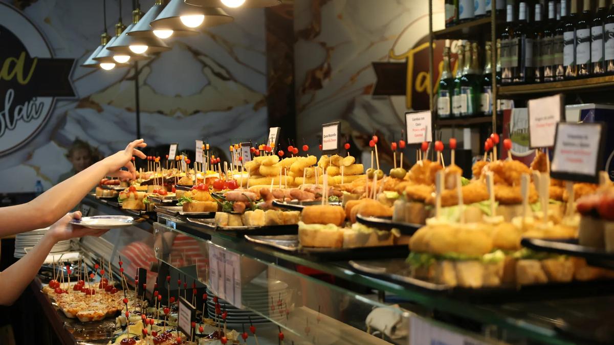 A variety of tapas and pintxos served on plates with toothpicks are displayed on a glass counter at a busy restaurant or bar, with people reaching for food.