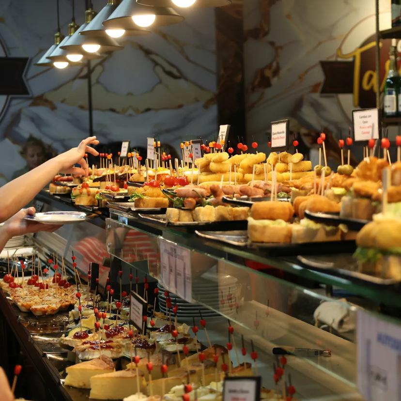 Auf einer Glasvitrine in einem gut besuchten Restaurant oder einer Bar sind verschiedene Tapas und Pintxos auf Tellern mit Zahnstochern ausgestellt, und die Gäste greifen nach den Speisen.