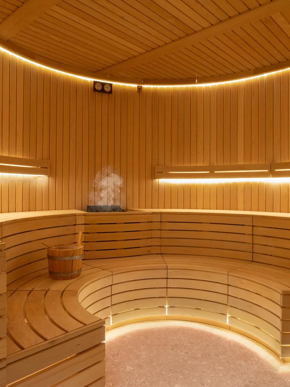 Modern wooden sauna with curved benches, warm yellow lighting, a wooden bucket, and a steaming stone heater in the center. The space appears clean, serene, and inviting for relaxation.