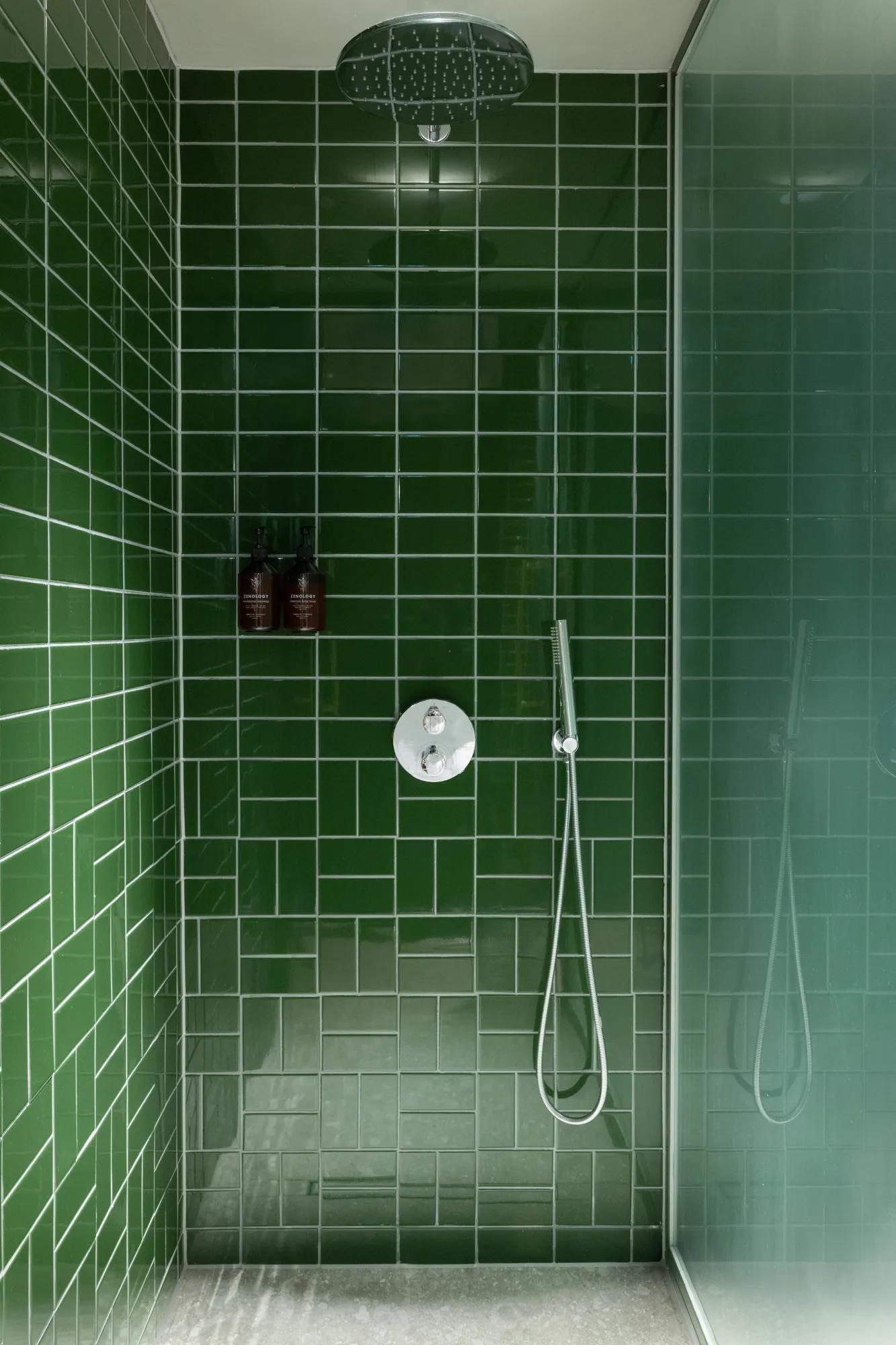 Eine moderne Dusche mit glänzenden grünen Metrofliesen, einem silbernen Duschkopf an der Decke, zwei Flaschen in einer Wandnische und einer an der Wand befestigten Handbrause. Auf der rechten Seite befindet sich eine Duschwand aus klarem Glas.