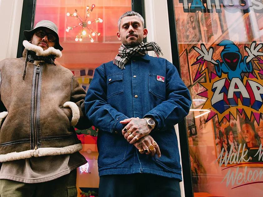 Zwei Männer stehen vor einem Schaufenster, das mit Comic-Aufklebern und Schildern im Graffiti-Stil verziert ist.