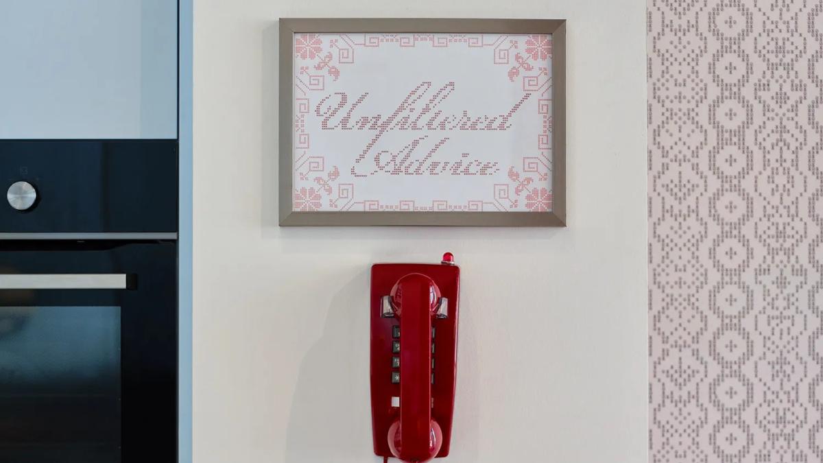 A red rotary phone is mounted on a wall beneath a framed sign that reads Unfiltered Advice. The wall features pastel tiles and part of a kitchen oven and towel are visible on the left.