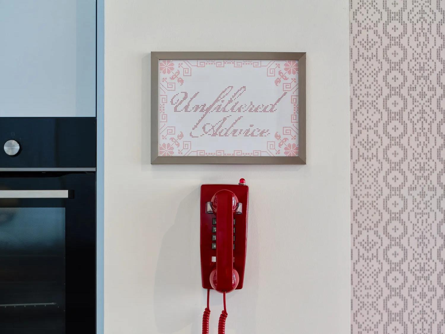 Un téléphone à cadran rouge est accroché au mur, sous une pancarte encadrée sur laquelle on peut lire « Unfiltered Advice ». Le mur est recouvert de carreaux aux tons pastel ; on aperçoit à gauche une partie d'un four de cuisine et une serviette.