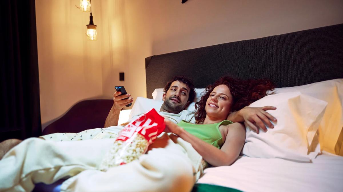 A couple relaxes in bed, smiling and watching TV together while sharing a bag of popcorn. The room is softly lit and cozy.