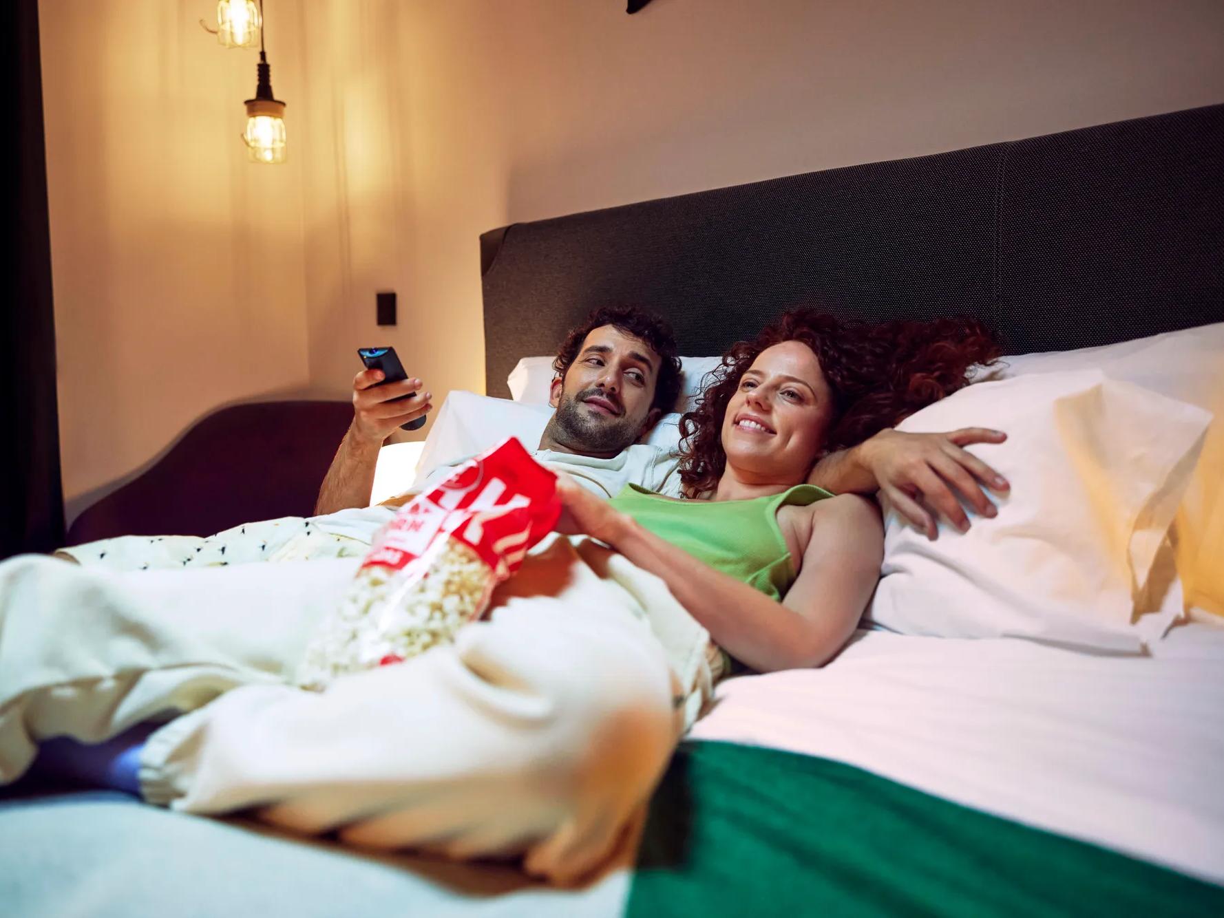 Una pareja se relaja en la cama, sonriendo y viendo la tele juntos mientras comparten una bolsa de palomitas. La habitación tiene una iluminación suave y resulta acogedora.