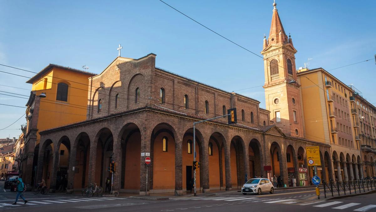A brick church with arched portico and a tall bell tower stands at a street corner in a city, with clear skies above and cars parked along the road.