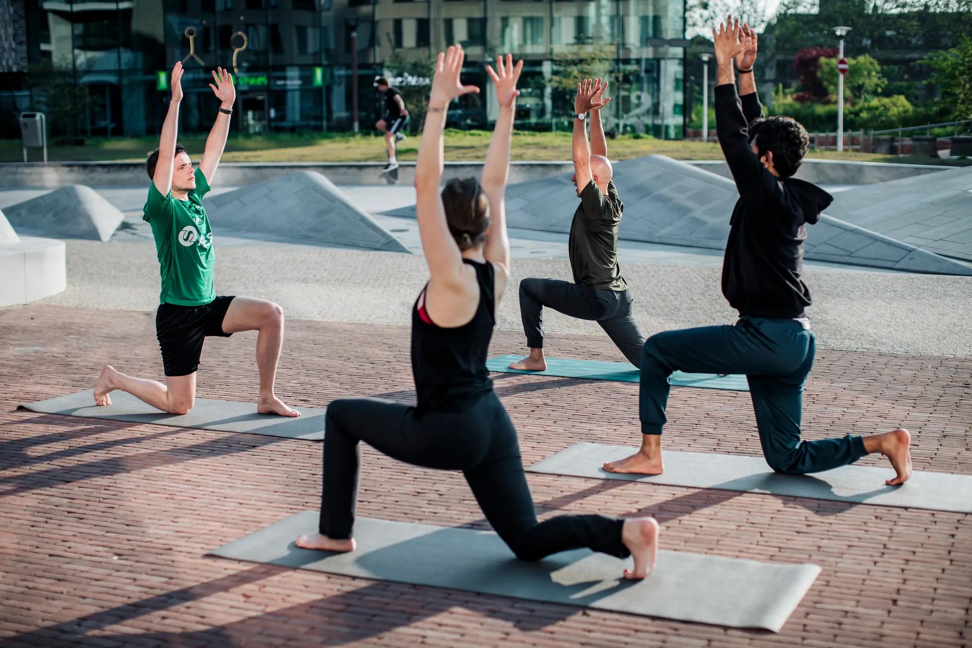 Four people practice yoga on mats outdoors, each in a kneeling lunge pose with arms raised overhead.