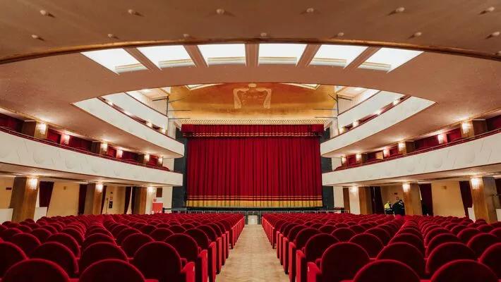 Rows of red seats face a stage with a large closed red curtain in an elegant theater auditorium with balconies and a decorative ceiling.