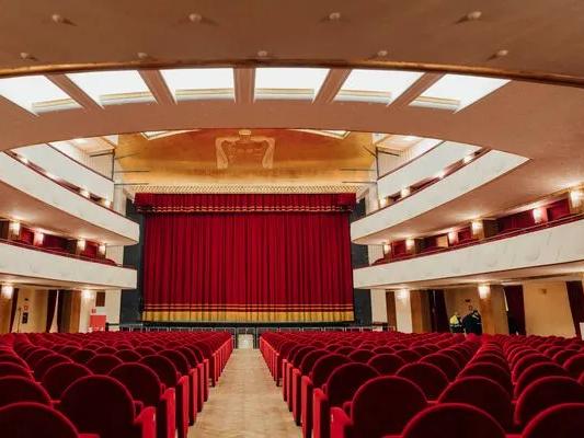 In einem eleganten Theatersaal mit Balkonen und einer verzierten Decke blicken Reihen roter Sitze auf eine Bühne, vor der ein großer, geschlossener roter Vorhang hängt.