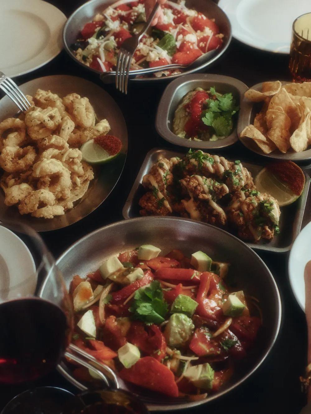 Four people sit around a table sharing plates of calamari, wings, chips with dip, salad, and glasses of red wine, preparing to enjoy a meal together.