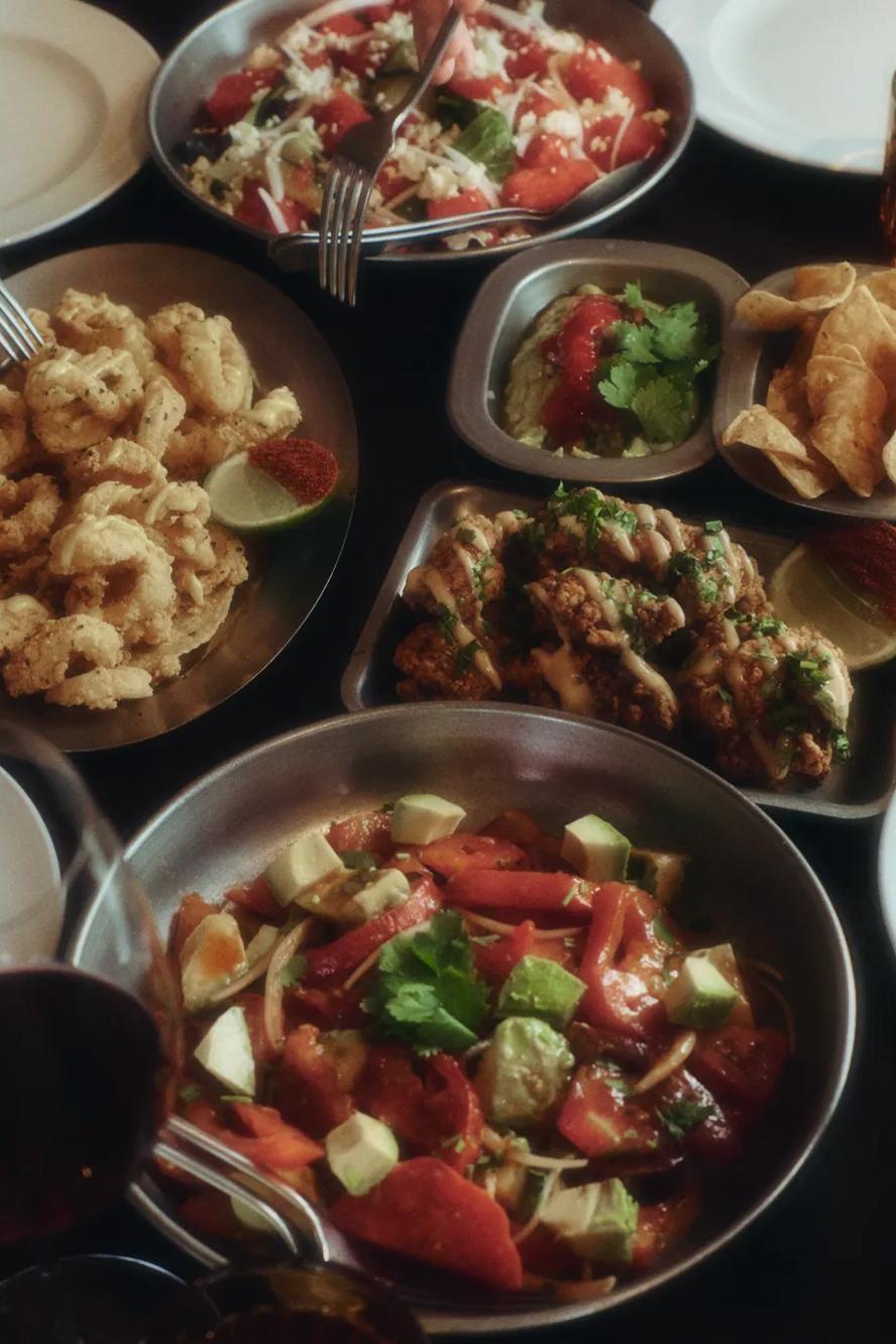 Four people sit around a table sharing plates of calamari, wings, chips with dip, salad, and glasses of red wine, preparing to enjoy a meal together.