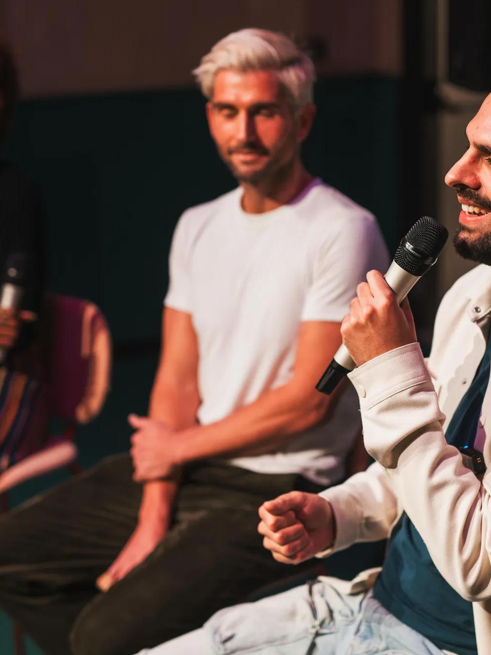 Four people sit on stage. One man in a light jacket speaks into a microphone, smiling. Another man in a white t-shirt and two women, one holding papers, listen. The setting appears informal and conversational.