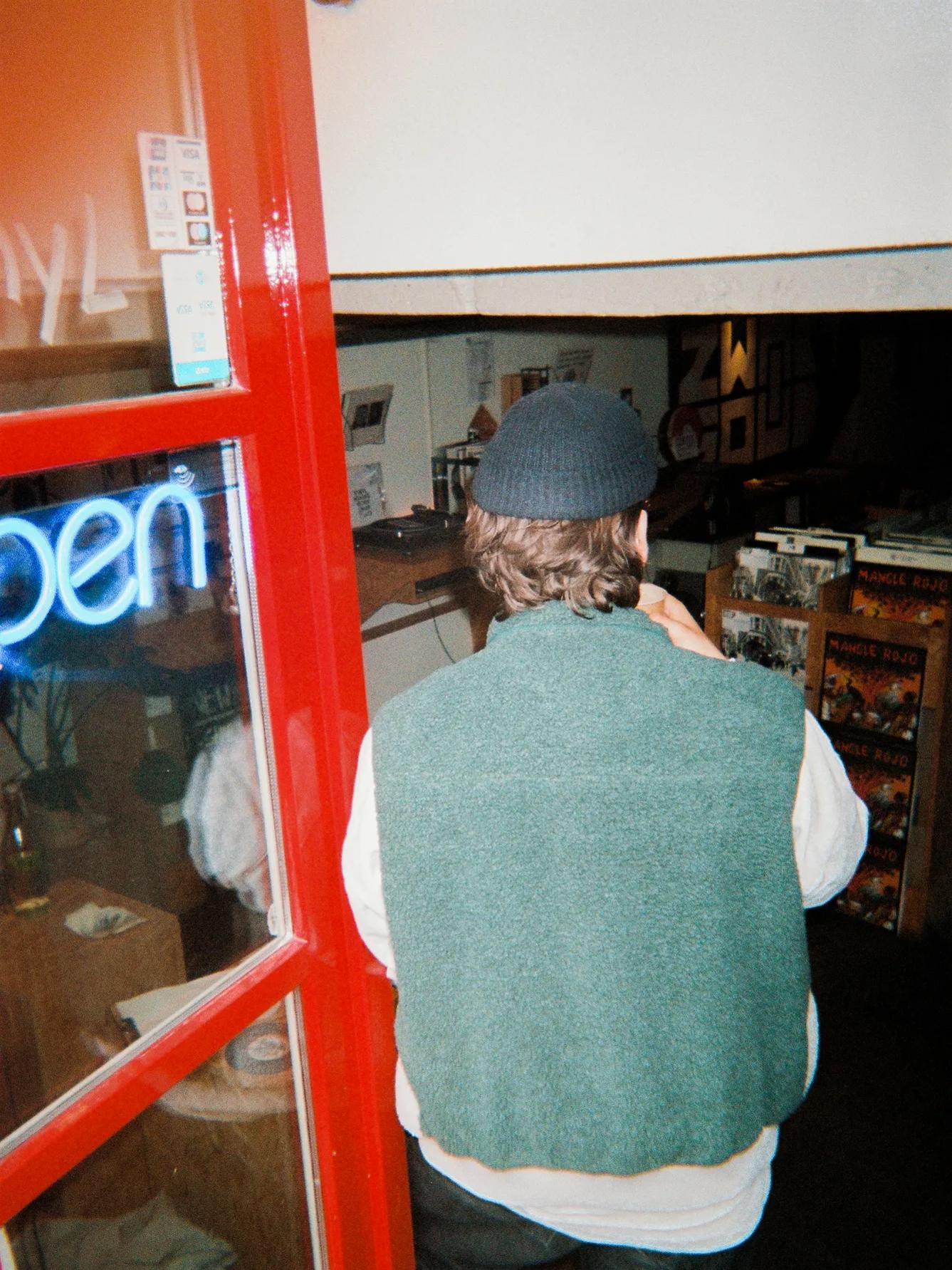 A person wearing a green vest and black beanie stands in the doorway of a shop with a neon open sign, facing inside.