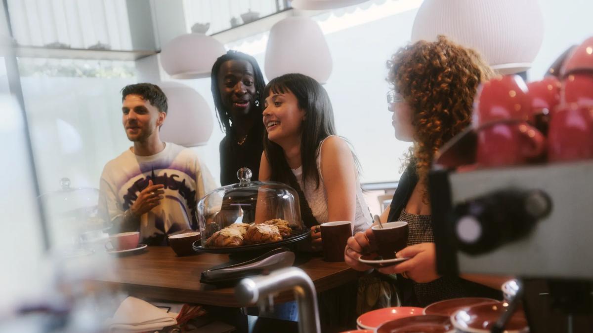 Four people sit at a cafe counter, smiling and talking. Coffee cups and pastries are on the counter, with stacked dishes in the foreground. The atmosphere appears relaxed and friendly.