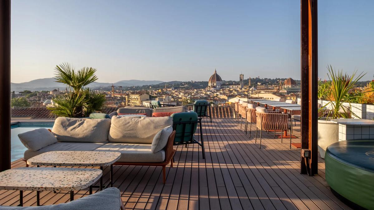 Rooftop lounge with modern seating, tables, and potted plants, overlooking Florences skyline with the Duomo and city buildings visible under a clear sky.
