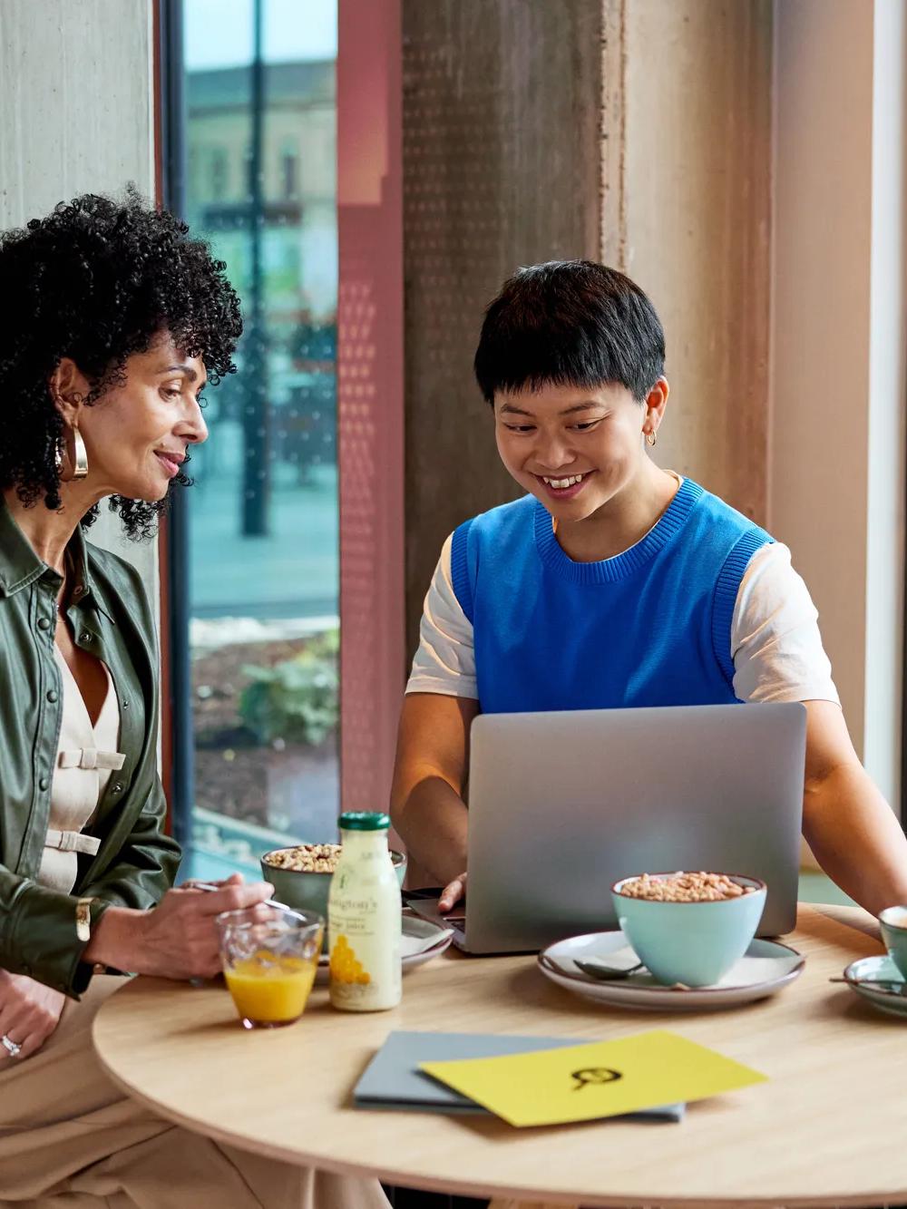 Two people sit at a round table with breakfast items, smiling and looking at a laptop. They are in a bright, modern cafe near large windows, with notebooks and drinks on the table.