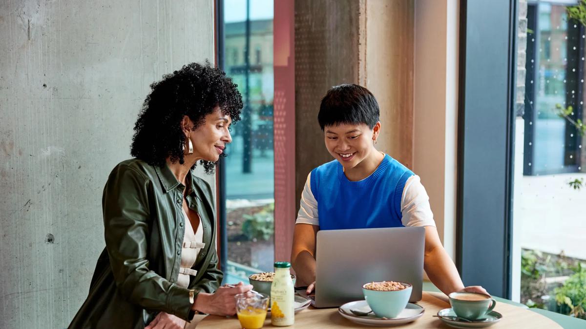 Two people sit at a round table with breakfast items, smiling and looking at a laptop. They are in a bright, modern cafe near large windows, with notebooks and drinks on the table.