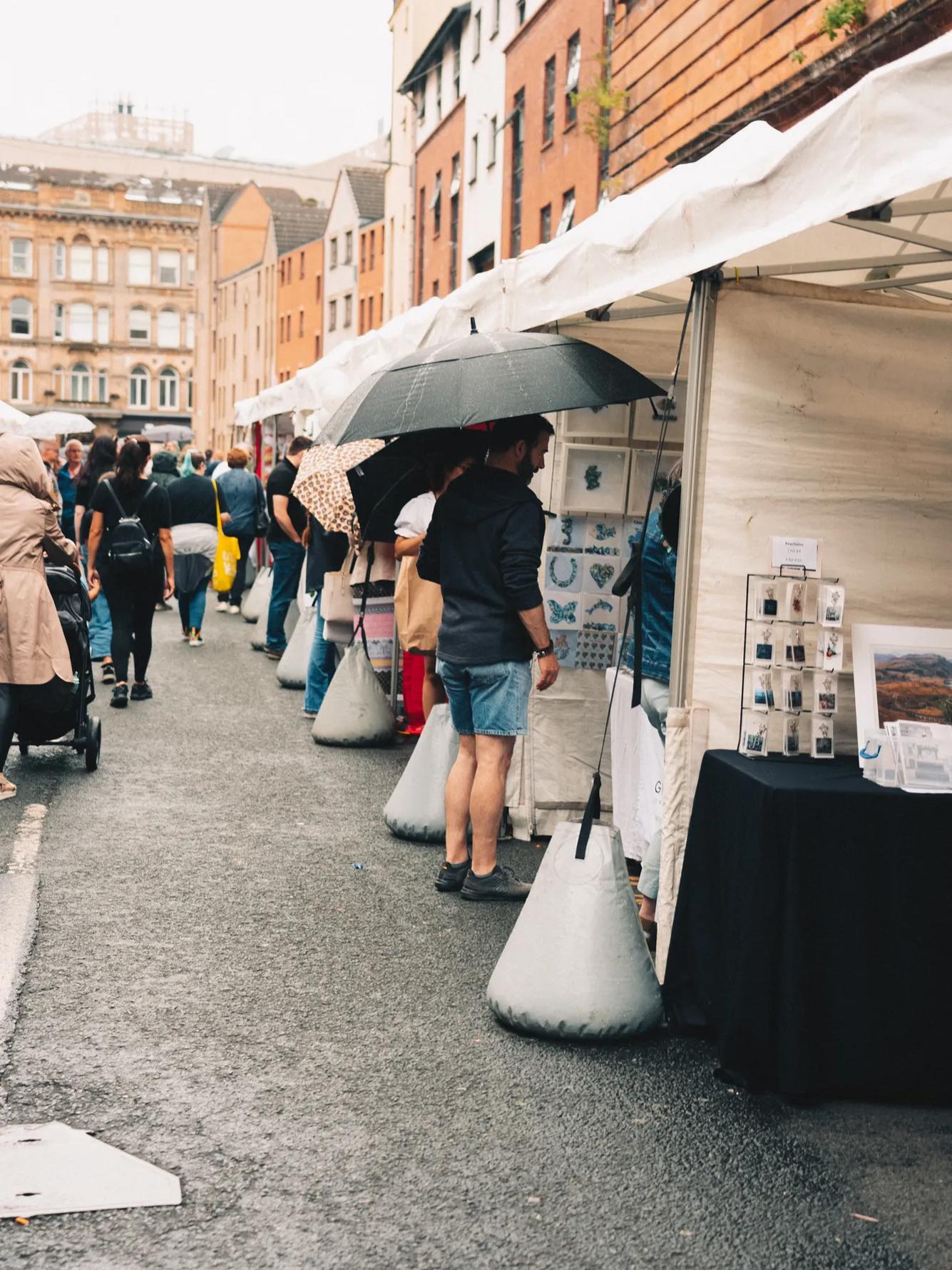 Outdoor market scene on a city street with people browsing stalls under white tents. Some shoppers hold umbrellas. Buildings line the background on a cloudy day.