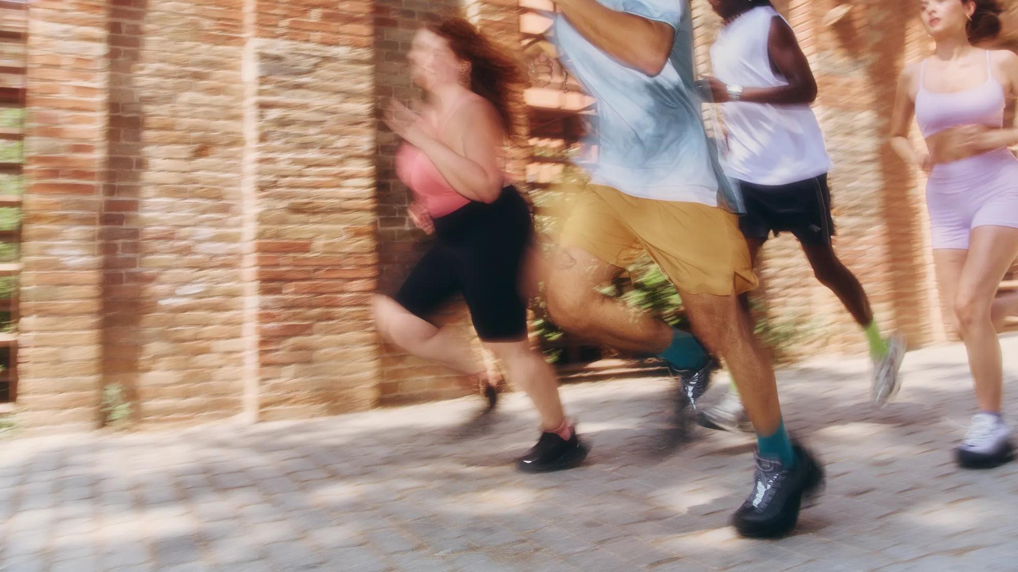 Four people are running outdoors beside a brick wall, captured in motion blur. The group includes men and women dressed in athletic wear, appearing to be exercising or racing together on a sunny day.