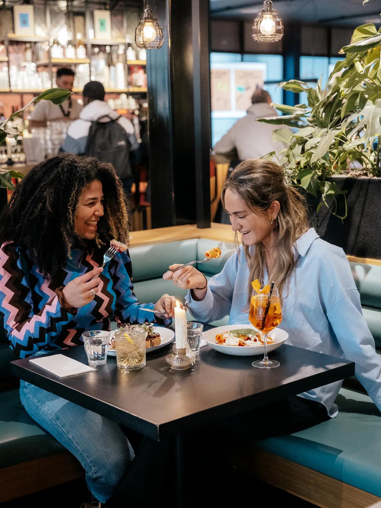 Two women sit at a restaurant booth, smiling and eating food. There are drinks, a lit candle, and plates on the table. The background shows other people, shelves, and plants in the cozy setting.