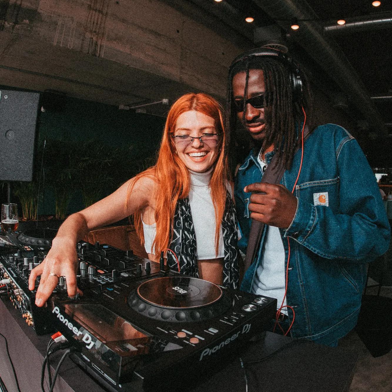 Two people are DJing together, smiling at a mixer. One wears glasses and a scarf, the other has headphones and a denim jacket. They seem to be enjoying the music in a lively indoor setting.
