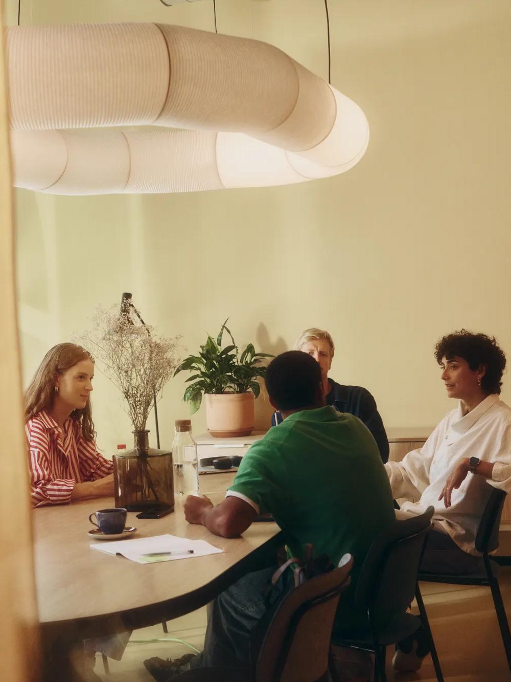 Four people sit around a wooden table in a warmly lit room, engaged in conversation. A plant and decorative vase are on the table and cabinet, with a large modern lamp hanging overhead.