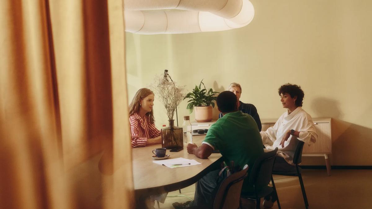 Quatre personnes sont assises autour d'une table en bois dans une pièce baignée d'une lumière chaleureuse, en pleine conversation. Une plante et un vase décoratif trônent sur la table et le meuble, tandis qu'une grande lampe moderne est suspendue au plafond.