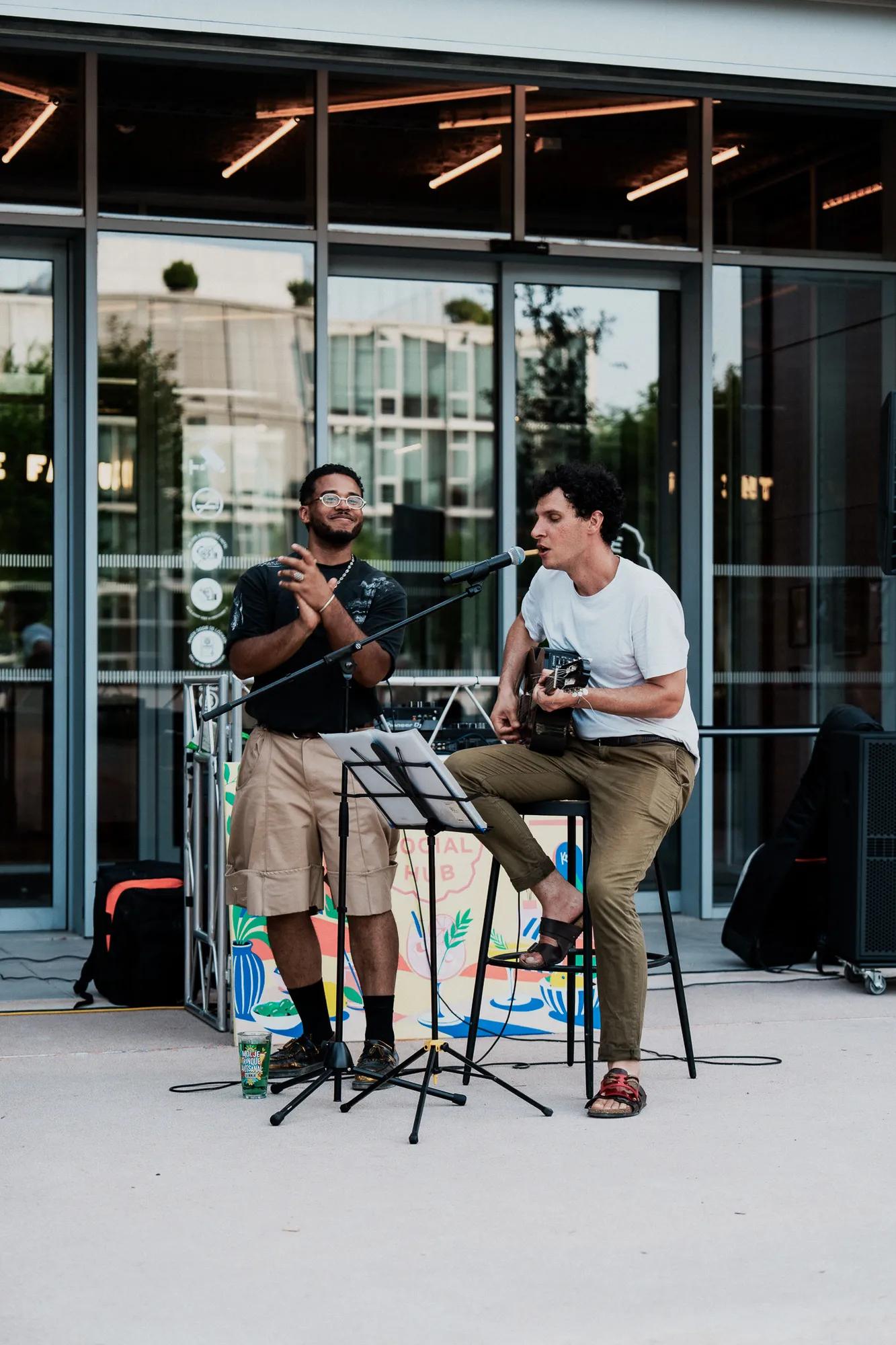 Two musicians perform outdoors. One plays guitar and sings into a microphone while seated; the other stands and claps. They are in front of large glass windows, with music equipment and decorations around them.