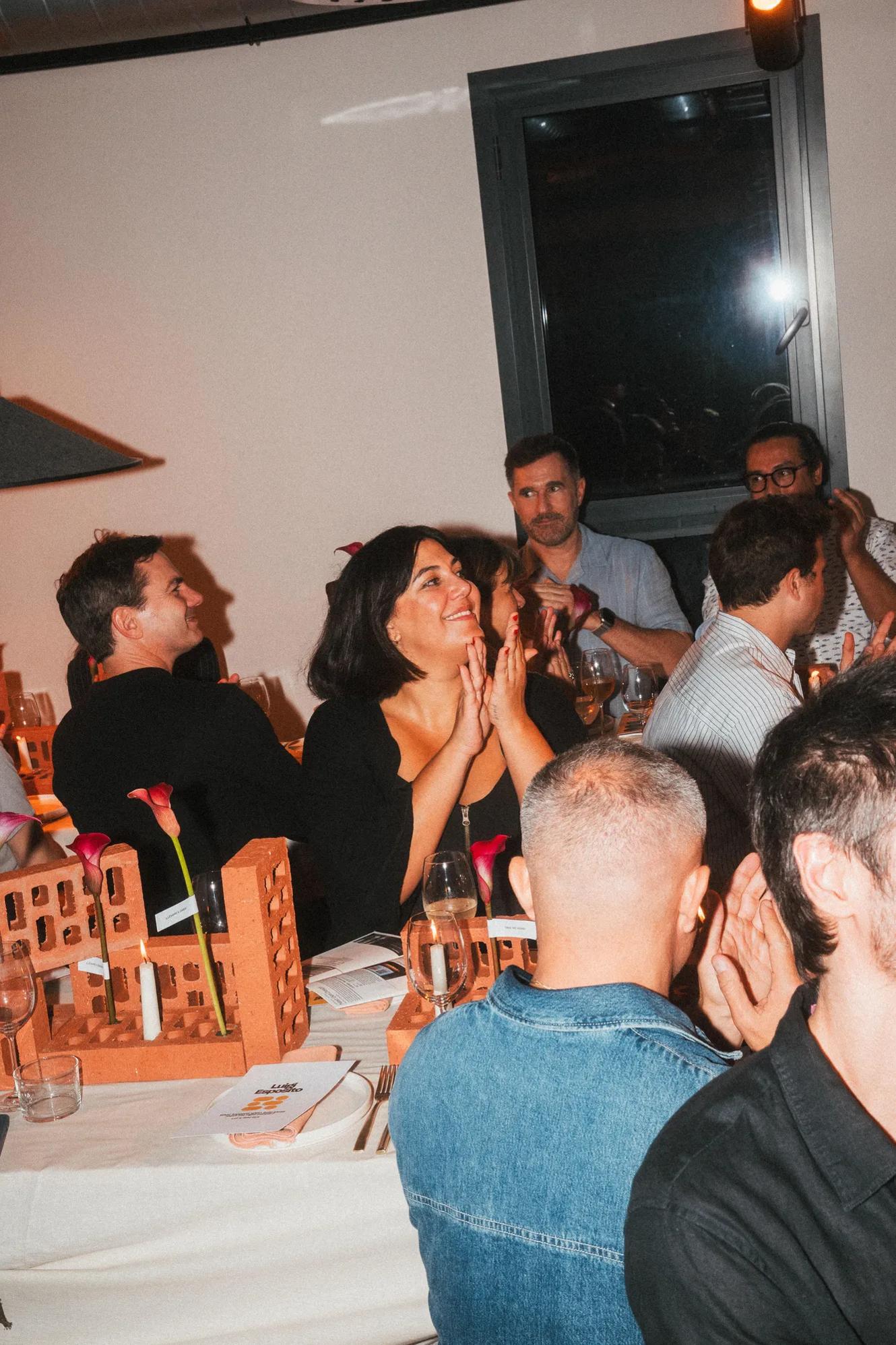 People seated at a dinner event, some applauding. Decorative wooden structures and glasses of wine are on the tables. The room has warm lighting and a window is visible in the background.