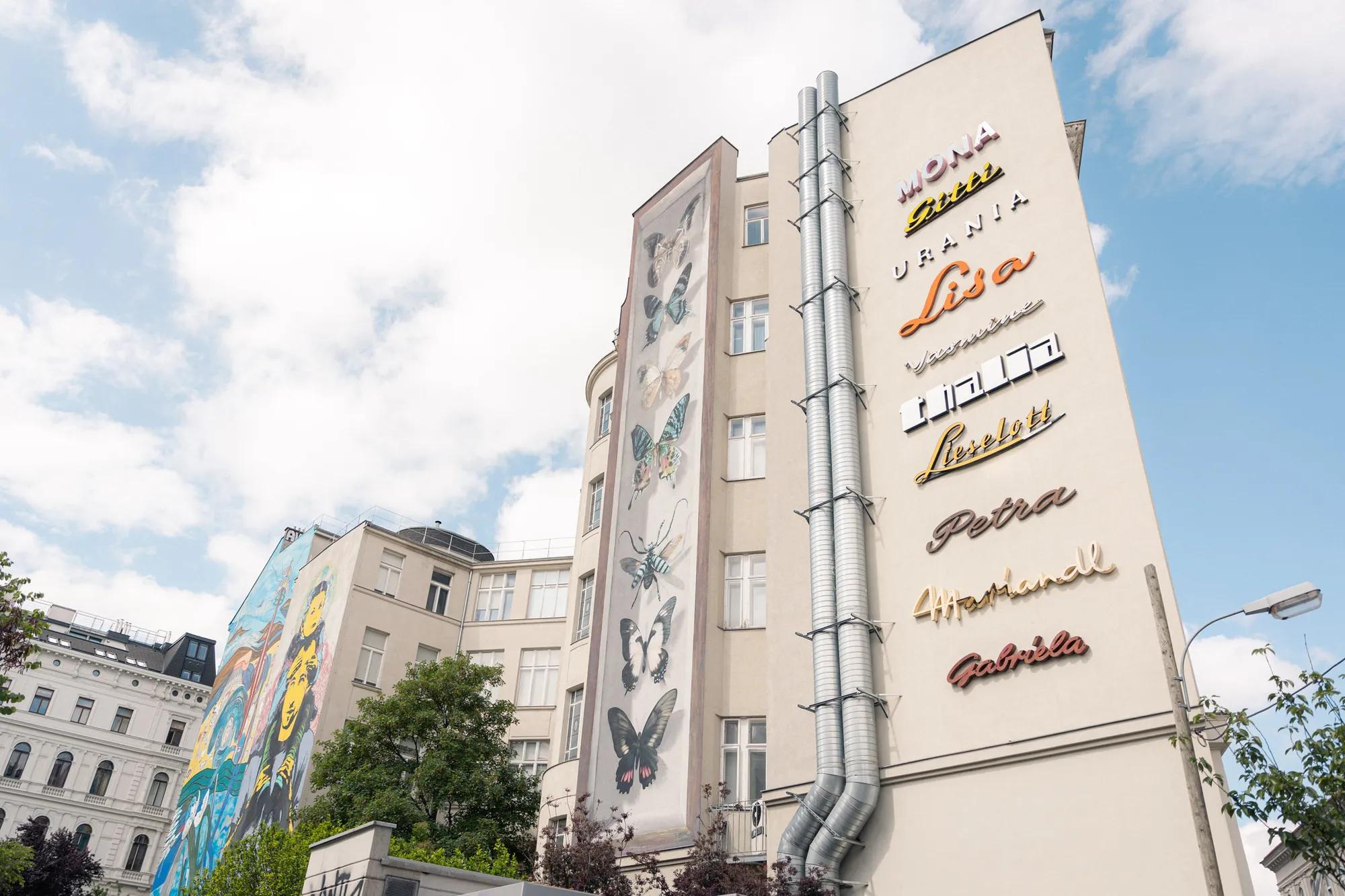 A white building with colorful names in neon lights on its wall, flanked by a mural with butterflies and another building featuring a vibrant, large-scale mural. The sky is partly cloudy.