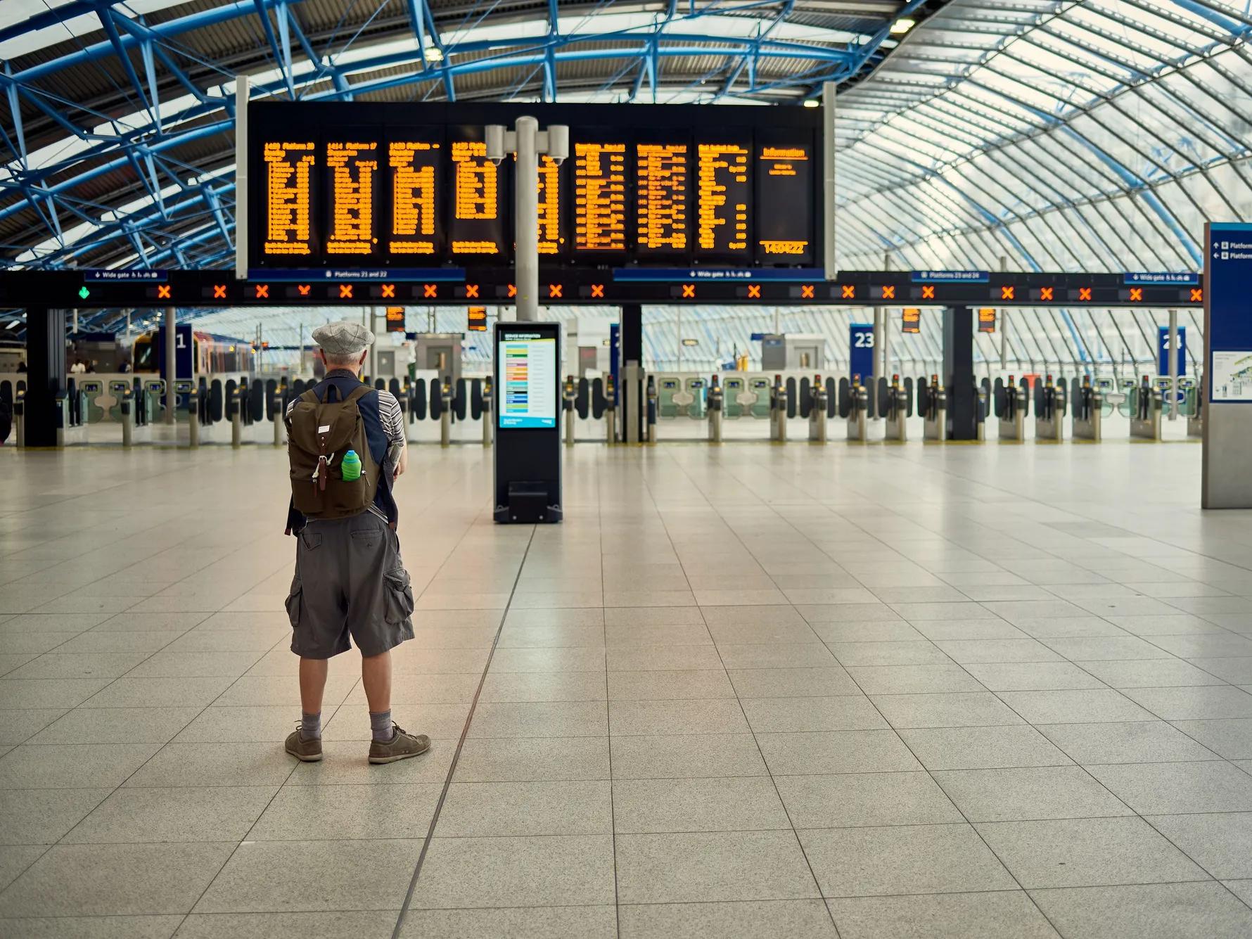Una persona con una mochila está de pie en una estación de tren espaciosa y moderna, mirando un gran panel digital de salidas en el que se muestran los horarios de los trenes. La estación es luminosa, con techos altos acristalados y un vestíbulo prácticamente vacío.