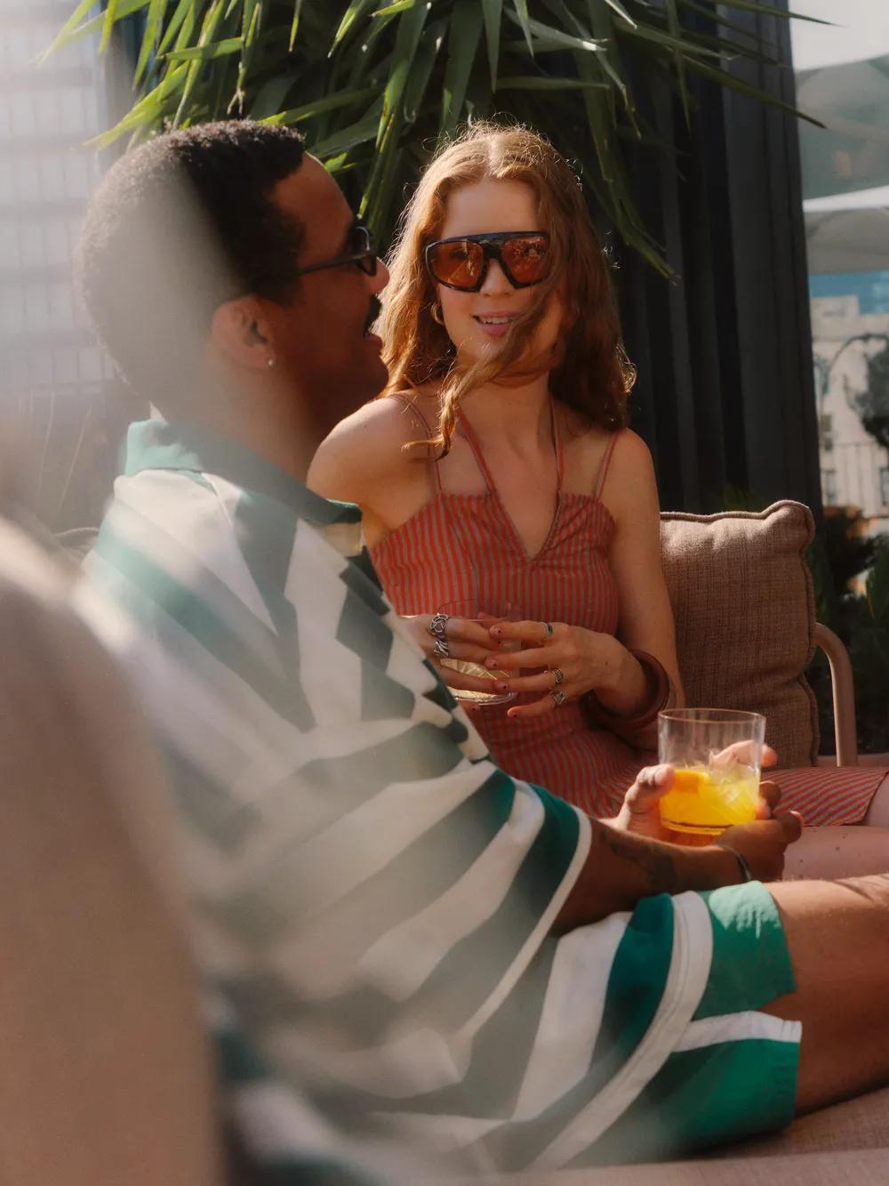 Two people relax outdoors on patio seating, chatting and holding drinks. One wears sunglasses and a dress, the other wears a striped shirt and shorts. Sunlight and plants create a warm, summery atmosphere.