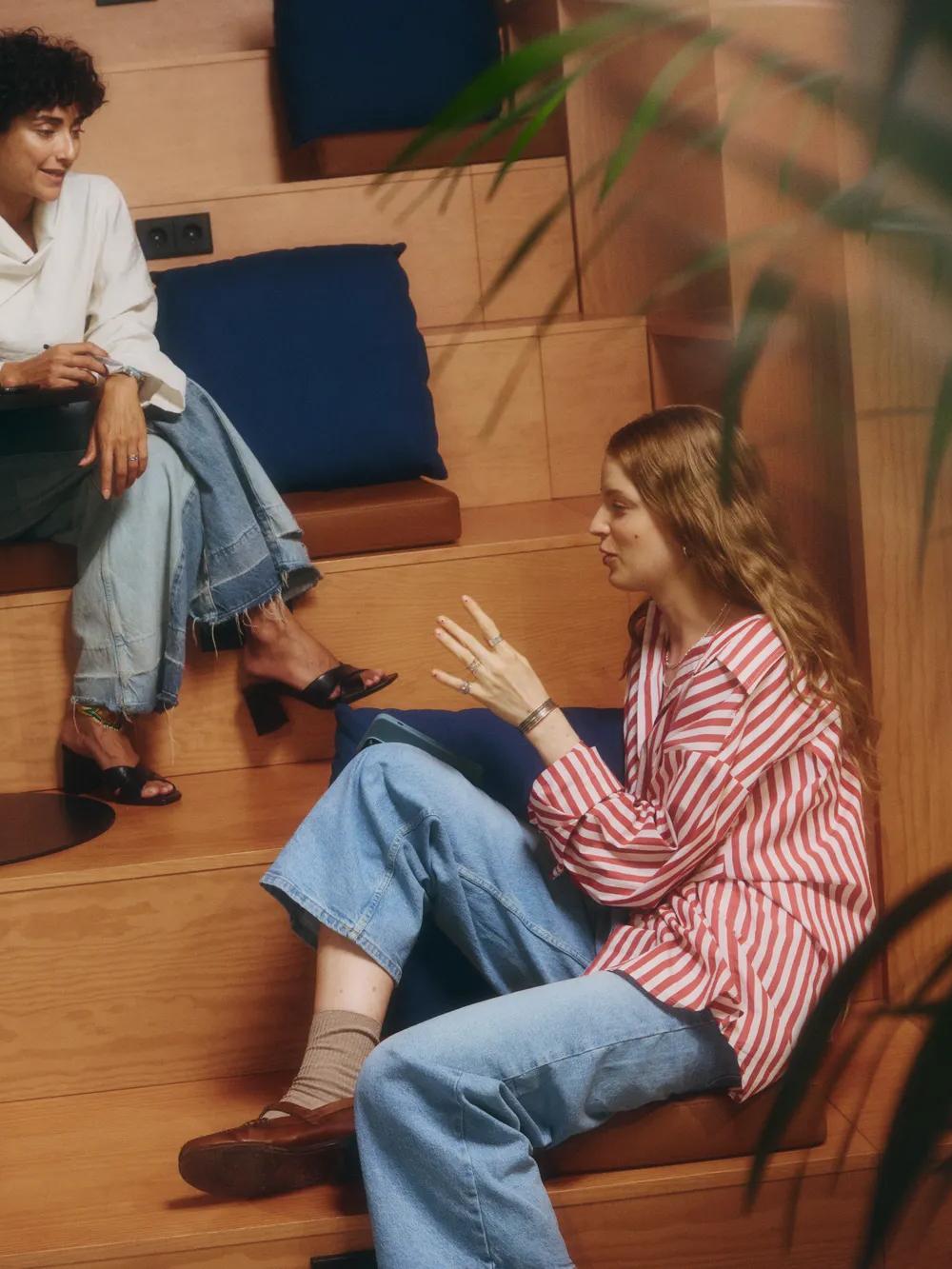 Two people sit on wooden steps with blue cushions, talking and smiling. One has a laptop on a small table. Blurred plants are in the foreground. Others are partially visible nearby. The atmosphere is casual and relaxed.