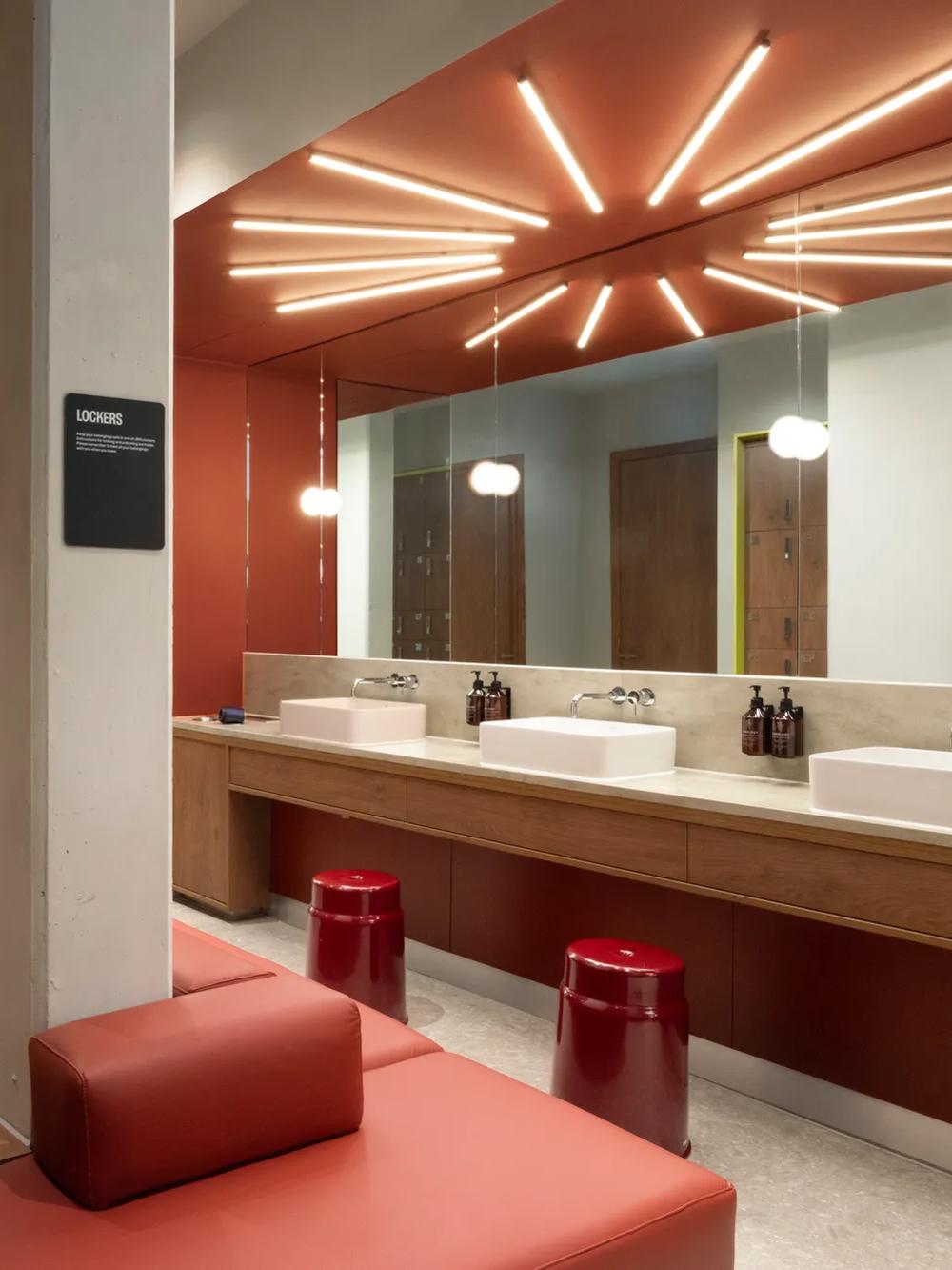 Modern restroom with red walls, long counter, two sinks, mirrors, red stools, and unique ceiling lights. Brown doors and cushioned seating are visible, creating a stylish, contemporary look.
