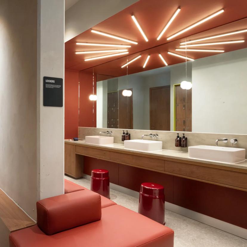Modern restroom with red walls, long counter, two sinks, mirrors, red stools, and unique ceiling lights. Brown doors and cushioned seating are visible, creating a stylish, contemporary look.
