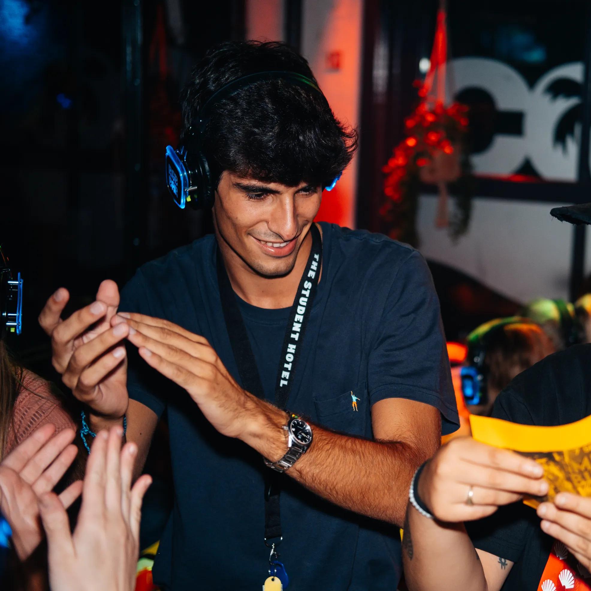 A young man wearing headphones smiles and dances with a crowd of people at a lively indoor event, illuminated by colorful lights.