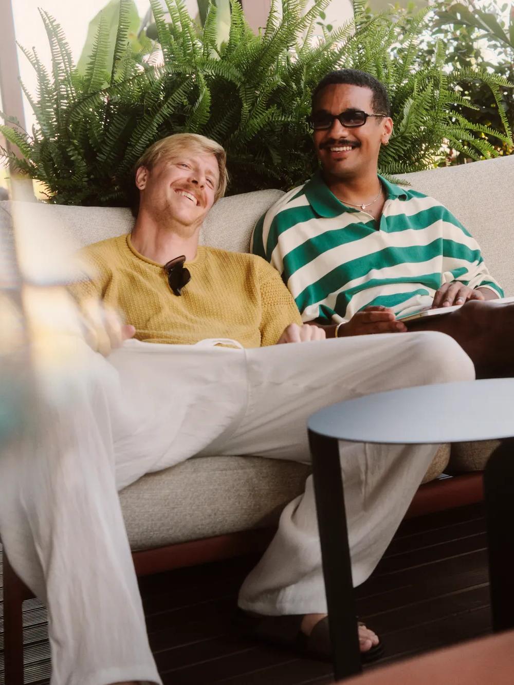 Two men sit relaxed on a sofa outdoors, smiling and talking with a third person in a purple top who is partially visible in the foreground. Green plants decorate the background.