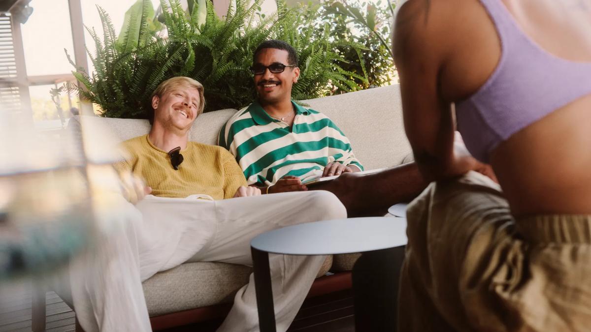 Two men sit relaxed on a sofa outdoors, smiling and talking with a third person in a purple top who is partially visible in the foreground. Green plants decorate the background.