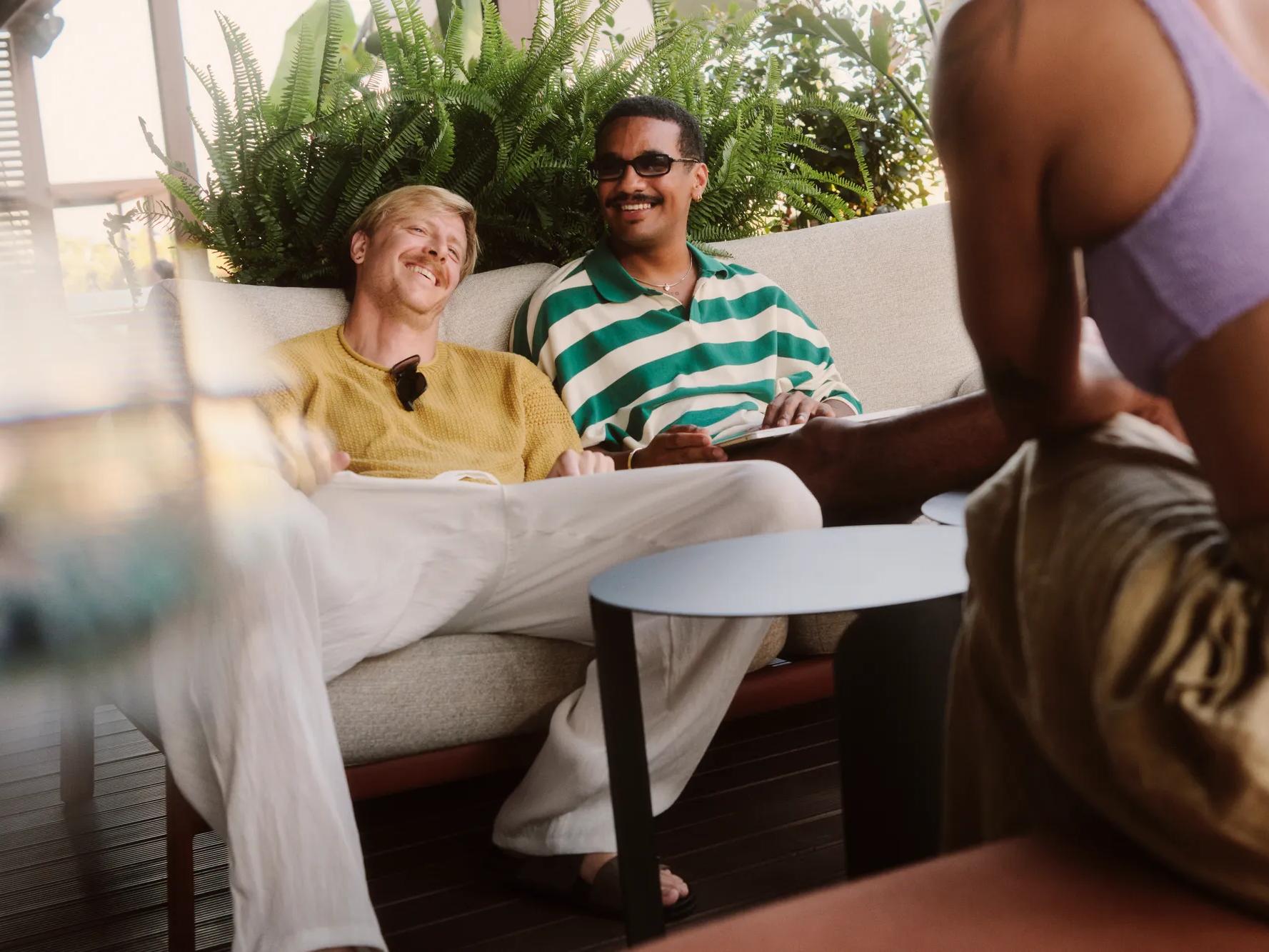 Dos hombres están sentados relajadamente en un sofá al aire libre, sonriendo y hablando con una tercera persona que lleva una camiseta morada y que se ve parcialmente en primer plano. El fondo está decorado con plantas verdes.