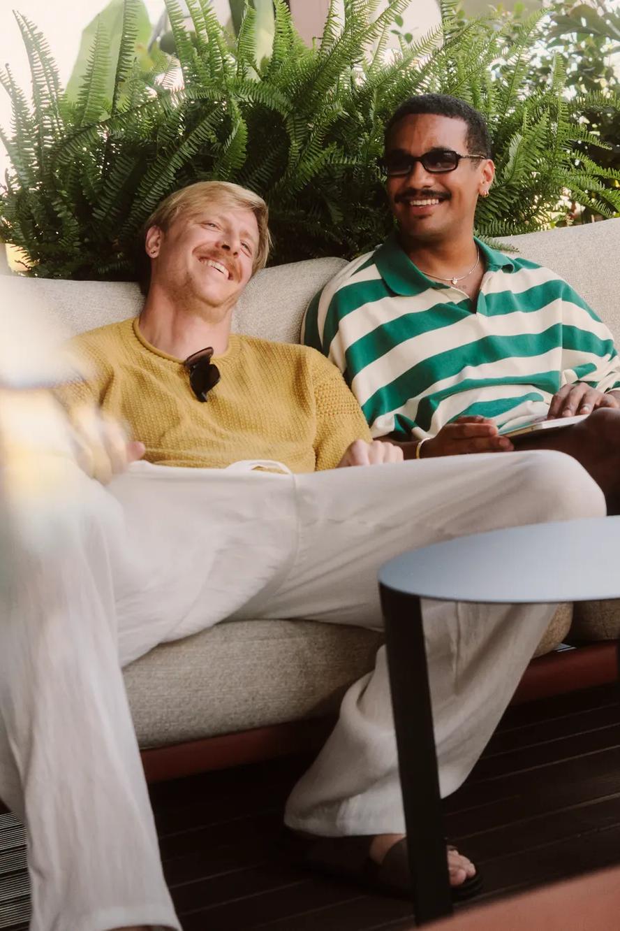 Two men sit relaxed on a sofa outdoors, smiling and talking with a third person in a purple top who is partially visible in the foreground. Green plants decorate the background.