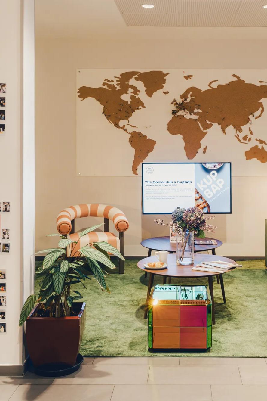 A cozy lounge with green sofa, striped chair, coffee table, and wall map. Polaroid photos are displayed on the left wall; plants and soft lighting create a welcoming atmosphere.