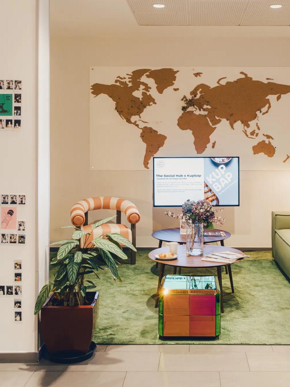 A cozy lounge with green sofa, striped chair, coffee table, and wall map. Polaroid photos are displayed on the left wall; plants and soft lighting create a welcoming atmosphere.